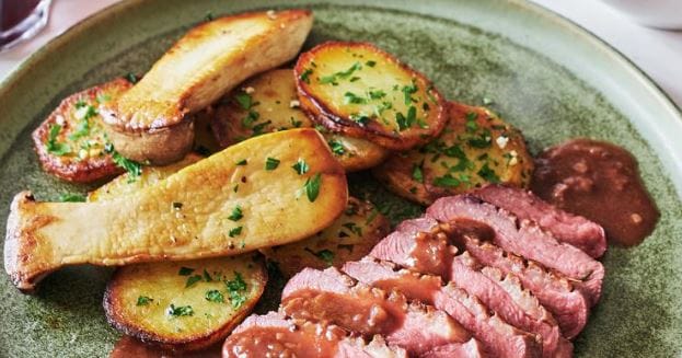 French-Malagasy fusion dish: sliced pink duck magret, accompanied by golden Sarladaise potatoes with parsley, a pan-fried king oyster mushroom, creamy brown sauce, on a green plate with a knife and a glass of red wine in the background.