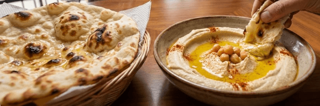 Main trempant un morceau de pain naan chaud dans un bol de houmous garni de pois chiches, d'huile d'olive et de paprika, servi sur une table en bois dans un restaurant en mezzanine avec vue sur la ville.